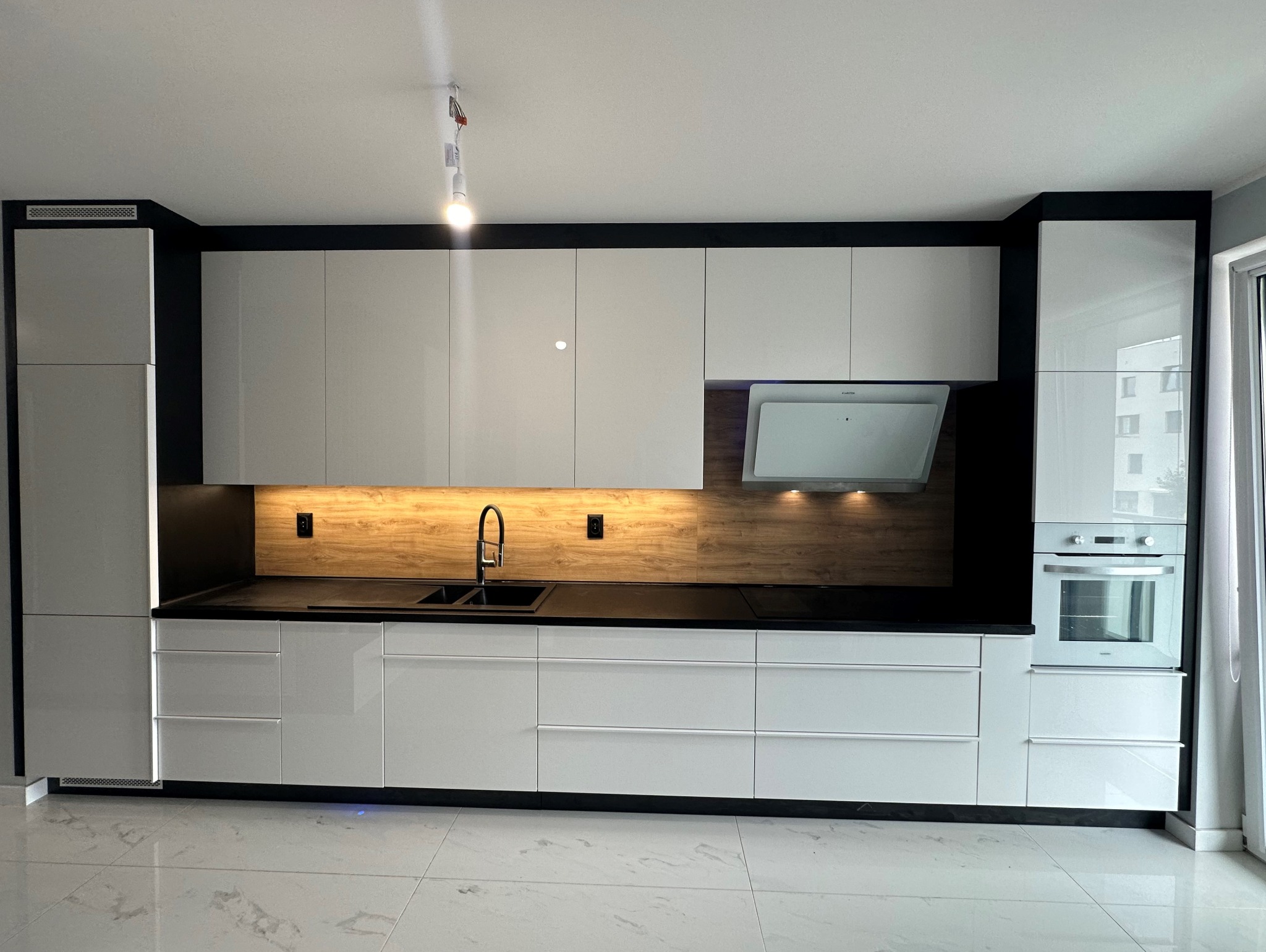 Nowoczesna, minimalistyczna kuchnia z białymi, lakierowanymi frontami szafek, czarnym blatem i drewnopodobną ścianą nad blatem, oświetlona punktowym światłem.