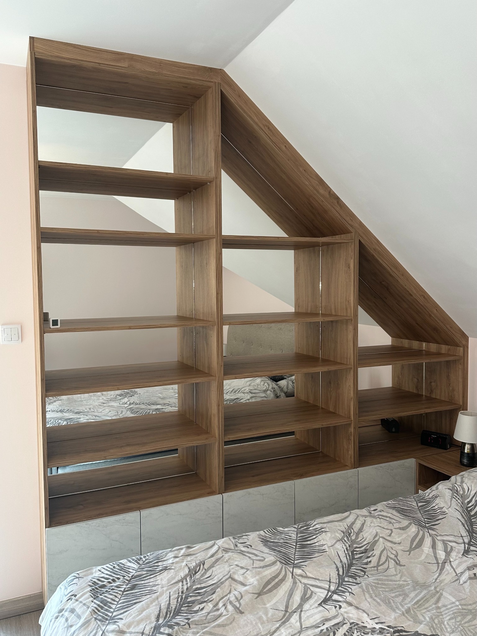 Drewniana szafa wnękowa z półkami, dopasowana do skosu dachu w sypialni, z szarymi frontami szafek dolnych i widoczną pościelą w liście.