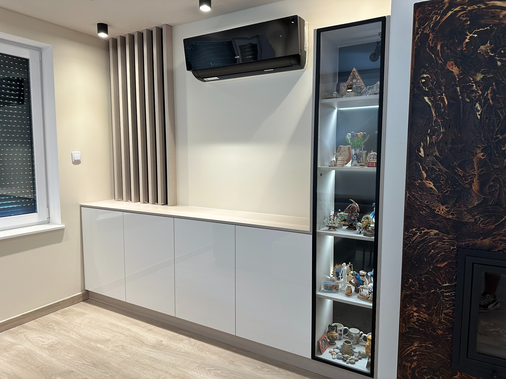 Nowoczesne meble do salonu z białymi, lakierowanymi frontami, szklana witryna z podświetleniem LED eksponująca dekoracyjne przedmioty, pionowe drewniane panele dekoracyjne na ścianie i klimatyzator.