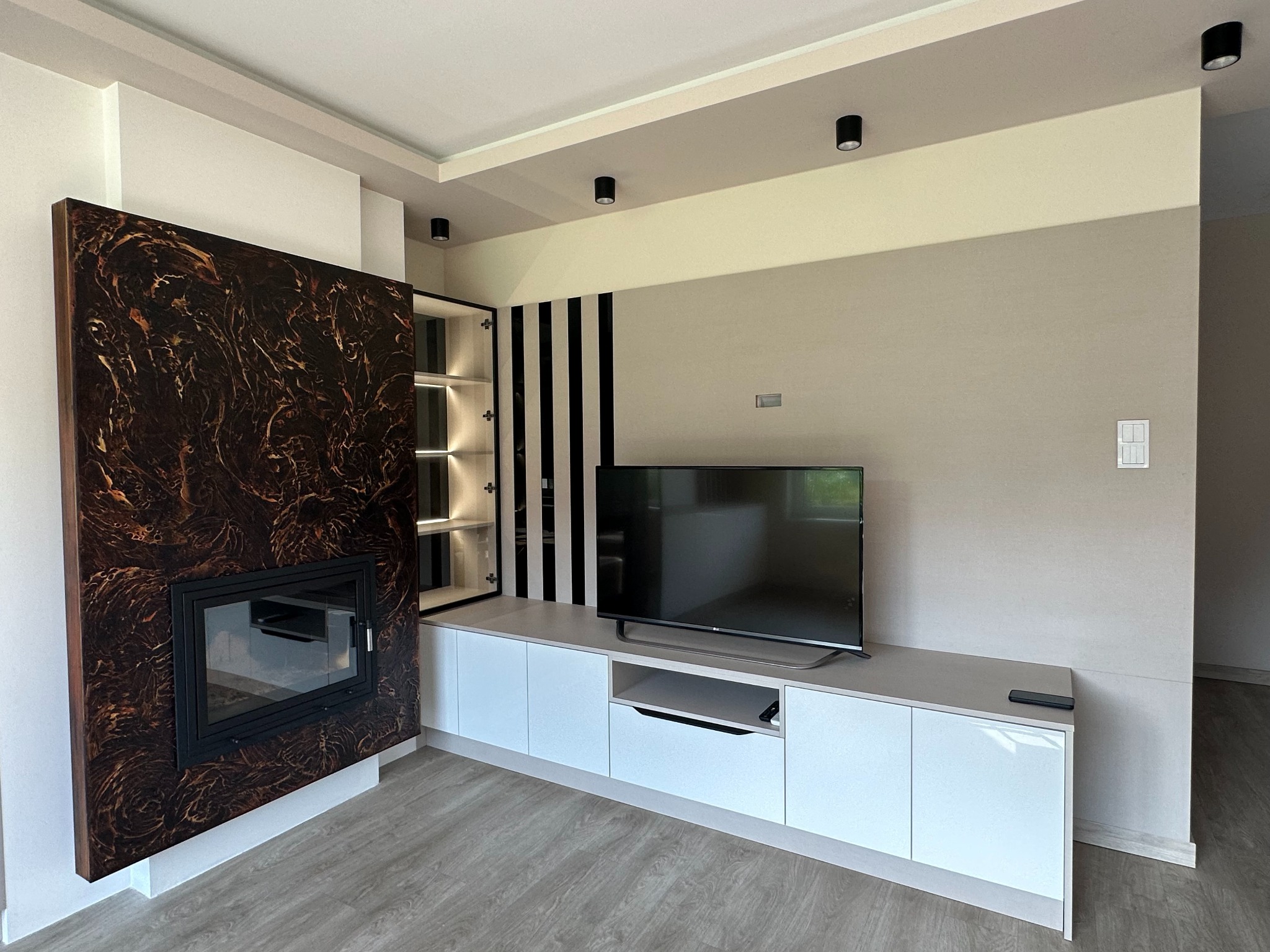 Nowoczesny salon z zabudową kominkową z ciemnego drewna o marmurkowym wzorze, białą szafką RTV z błyszczącymi frontami, telewizorem i czarnymi paskami dekoracyjnymi na ścianie.