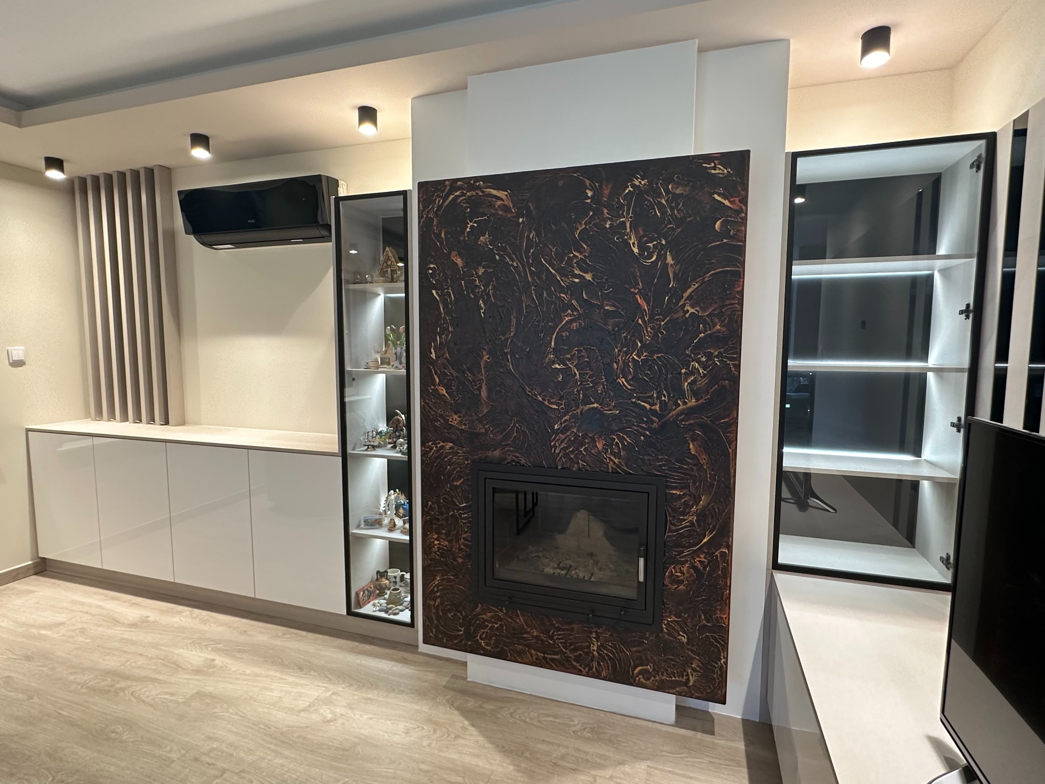 Nowoczesny salon z zabudową meblową w kolorze białym, zintegrowaną z kominkiem o dekoracyjnej obudowie w odcieniach brązu i złota, oraz przeszklonymi witrynami podświetlonymi LED.