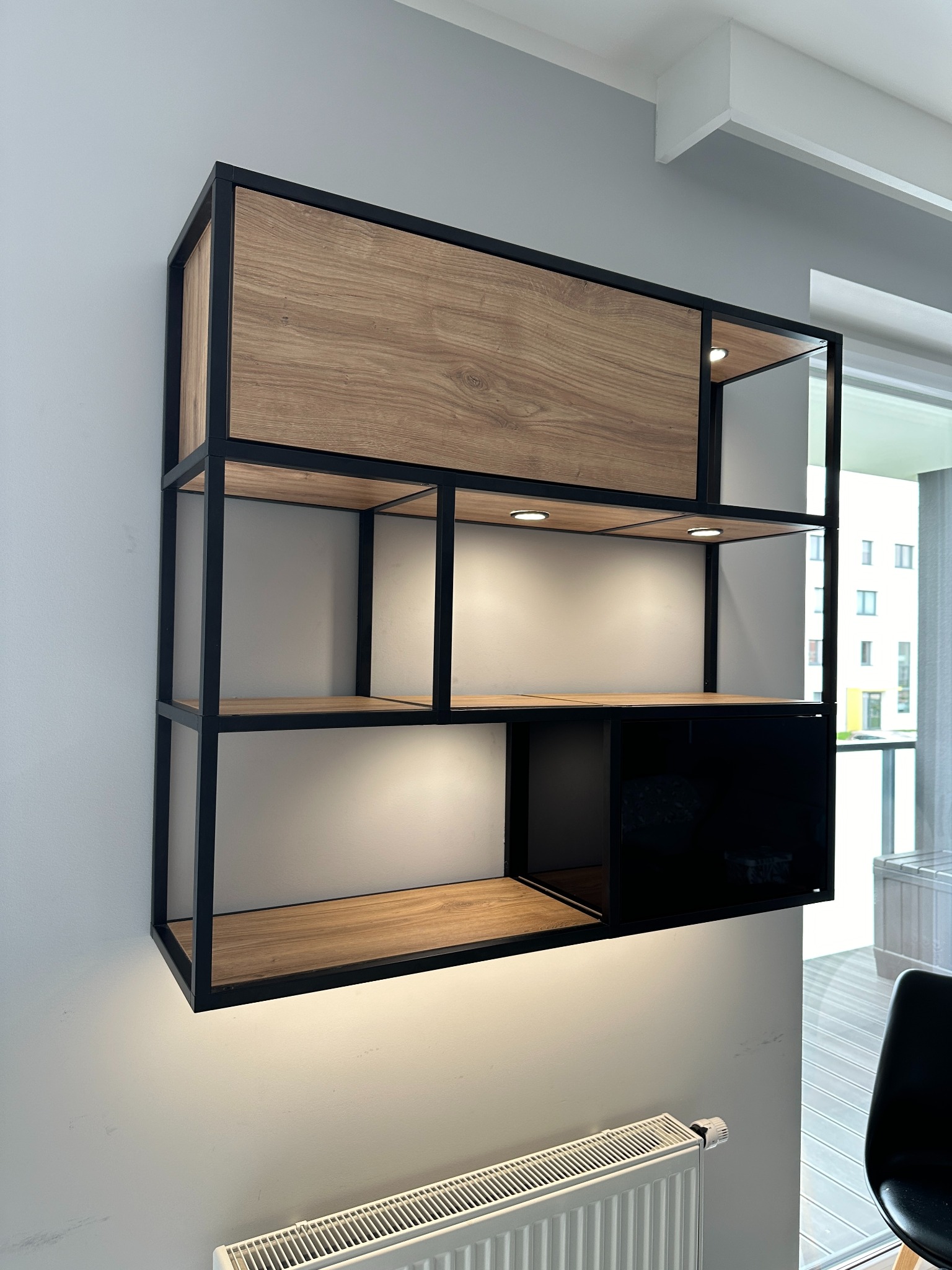 Półka wisząca z drewnianymi i czarnymi elementami, oświetleniem punktowym LED, zamontowana na szarej ścianie nad grzejnikiem.