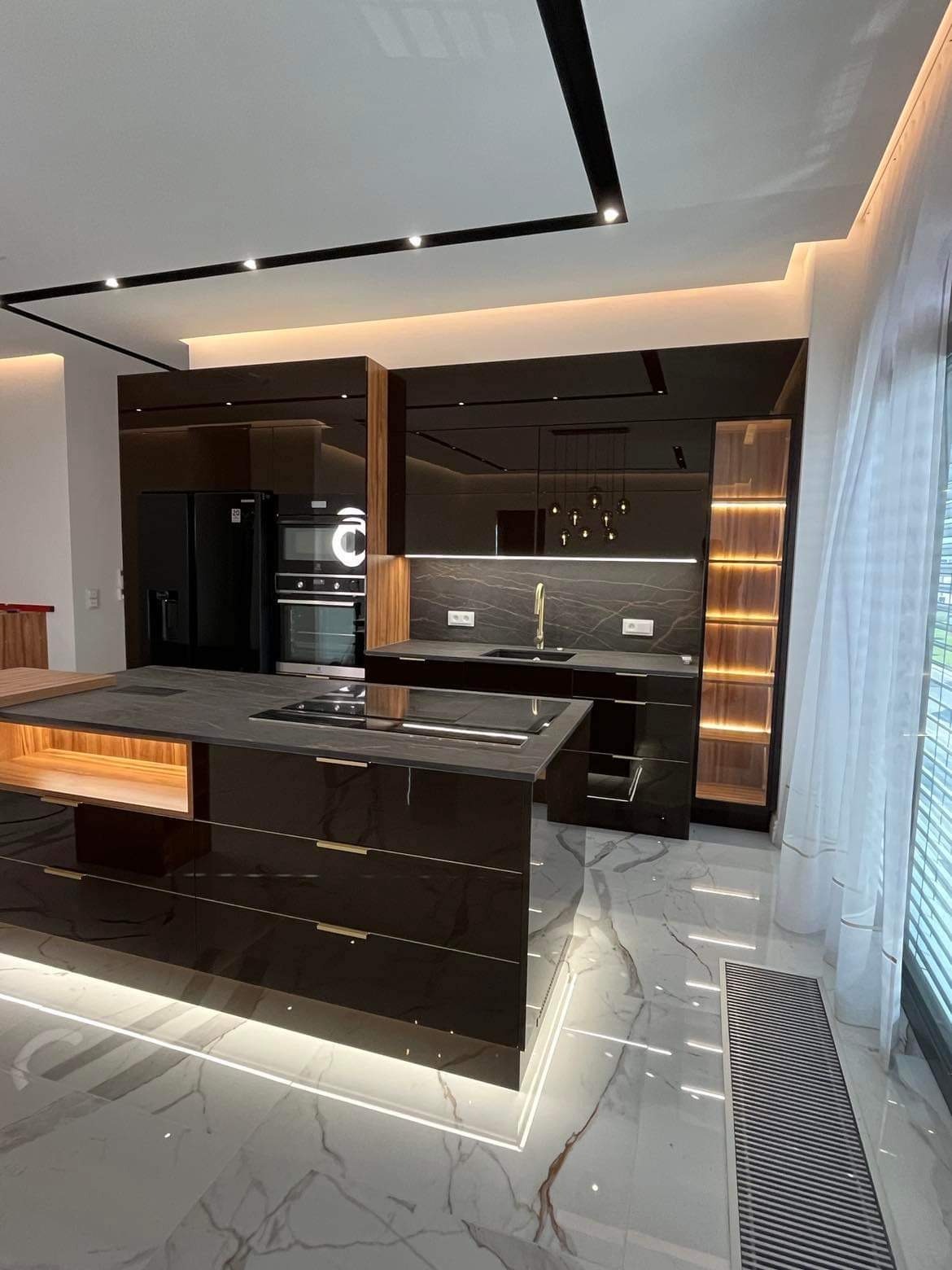 Nowoczesna, czarna kuchnia z połyskiem, wyspą z blatem imitującym kamień, oświetleniem LED pod szafkami i marmurową podłogą.