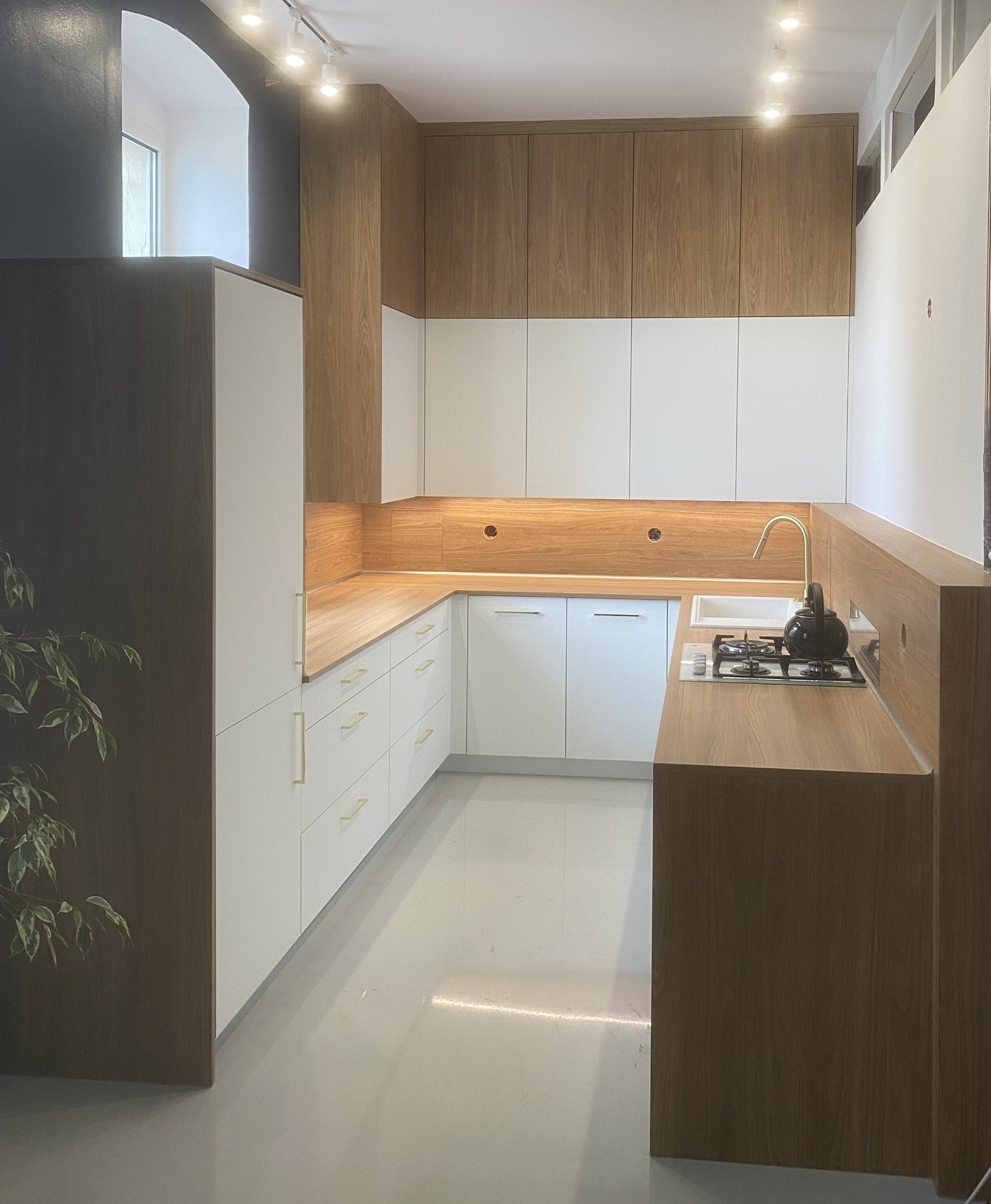 Nowoczesna kuchnia z białymi frontami i drewnianymi blatami, złote uchwyty, czajnik na płycie grzewczej, oświetlenie punktowe, minimalistyczny design.