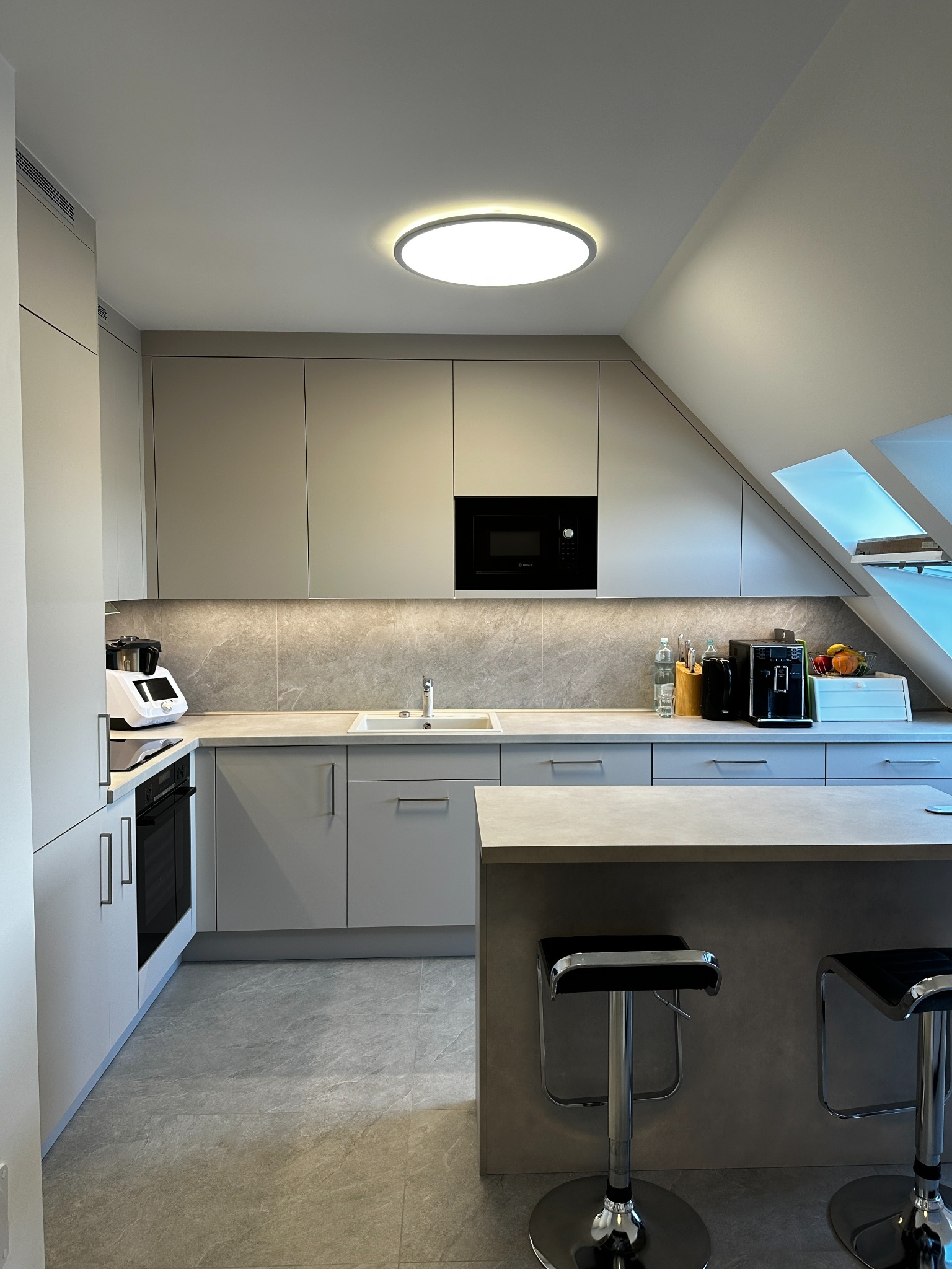 Jasna kuchnia na poddaszu z wyspą i hokerami. Zabudowa w odcieniach szarości, okap wbudowany, oświetlenie LED pod szafkami. Widoczne okna dachowe.