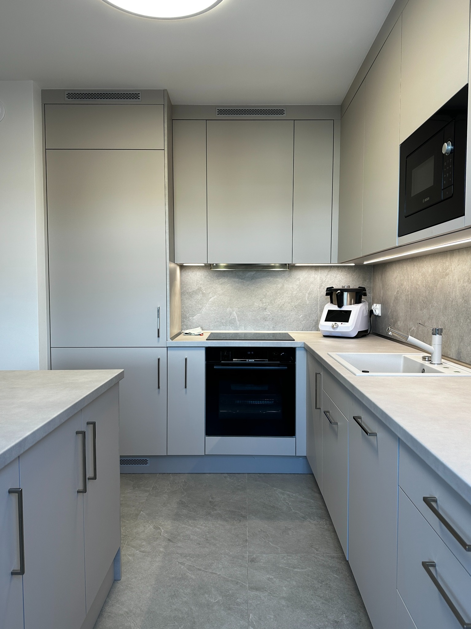 Nowoczesna kuchnia w odcieniach szarości z subtelnym oświetleniem LED, z piekarnikiem Bosch i marmurowym blatem oraz sprzętem AGD na blacie.