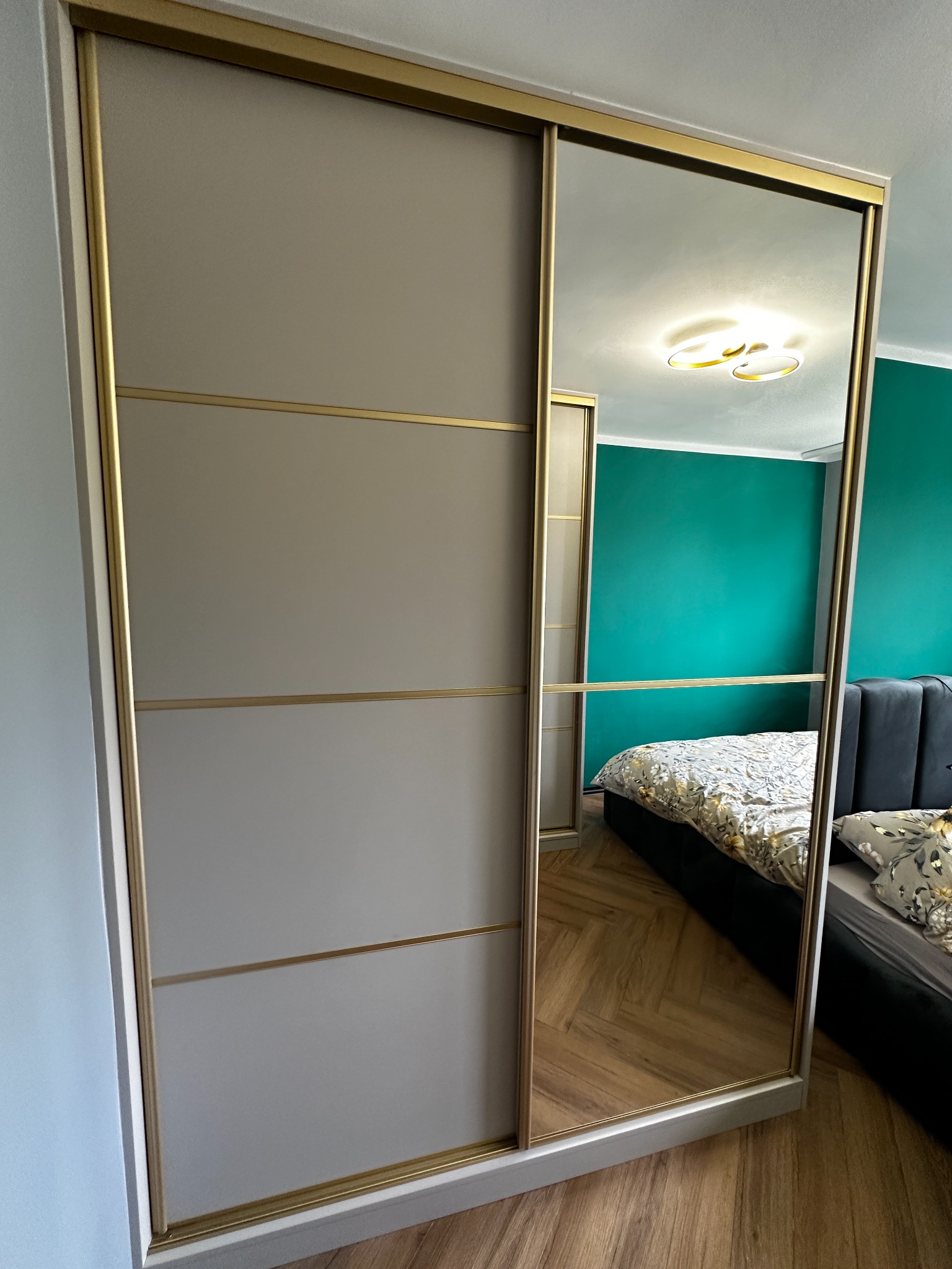 Szafa wnękowa z przesuwnymi drzwiami, beżowe fronty z złotymi akcentami. W lustrzanych drzwiach odbija się wnętrze sypialni z zieloną ścianą i łóżkiem.
