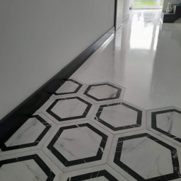 OSKAR Mariusz Kmiecik - Biała podłoga z połyskiem, przechodząca w heksagonalne płytki imitujące marmur z czarnymi obwódkami, widoczna listwa przypodłogowa i fragment okna z widokiem na zewnątrz.