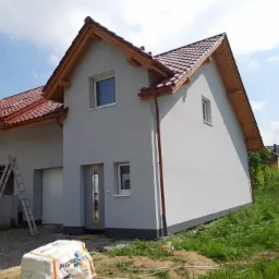 Świeżo pomalowany dom jednorodzinny w odcieniach szarości z czerwoną dachówką, widoczna drabina oparta o ścianę i kostka styropianu na trawie.