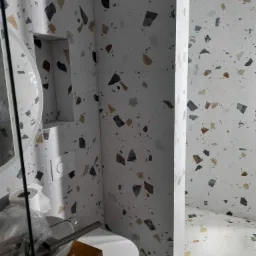 Biała łazienka wykończona płytkami imitującymi lastryko z nieregularnymi, kolorowymi fragmentami kamieni, widoczna umywalka z akcesoriami i toaleta