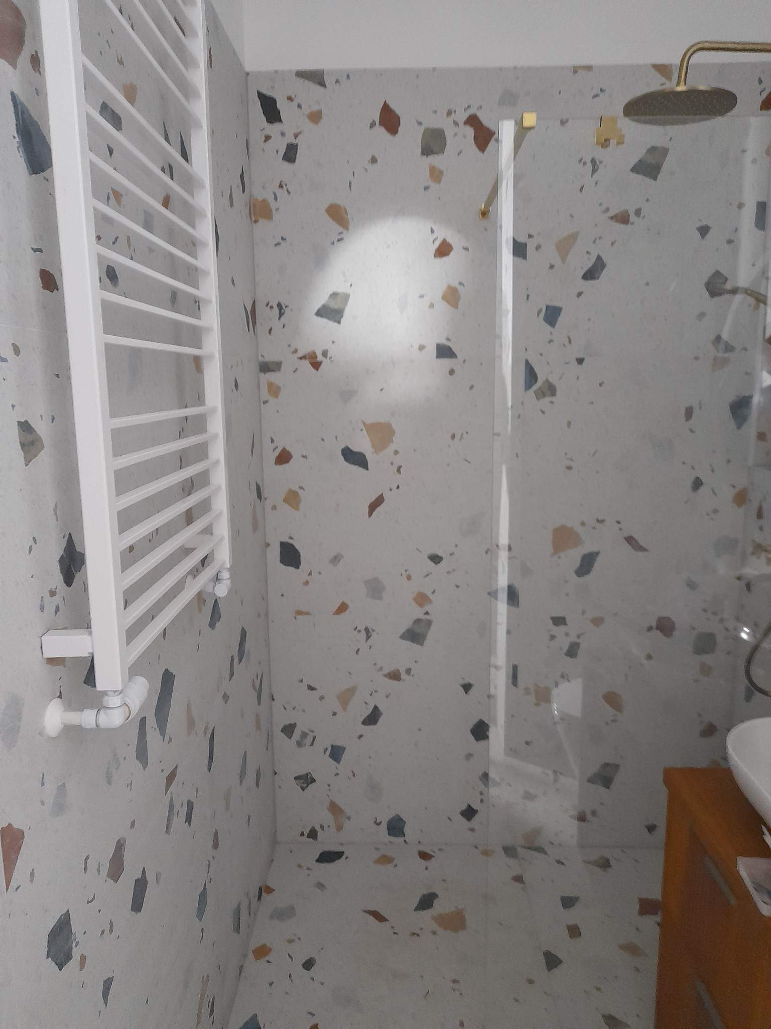 Wnętrze łazienki z kabiną prysznicową typu walk-in, grzejnikiem drabinkowym i umywalką na drewnianej szafce, wykończone płytkami imitującymi lastryko.