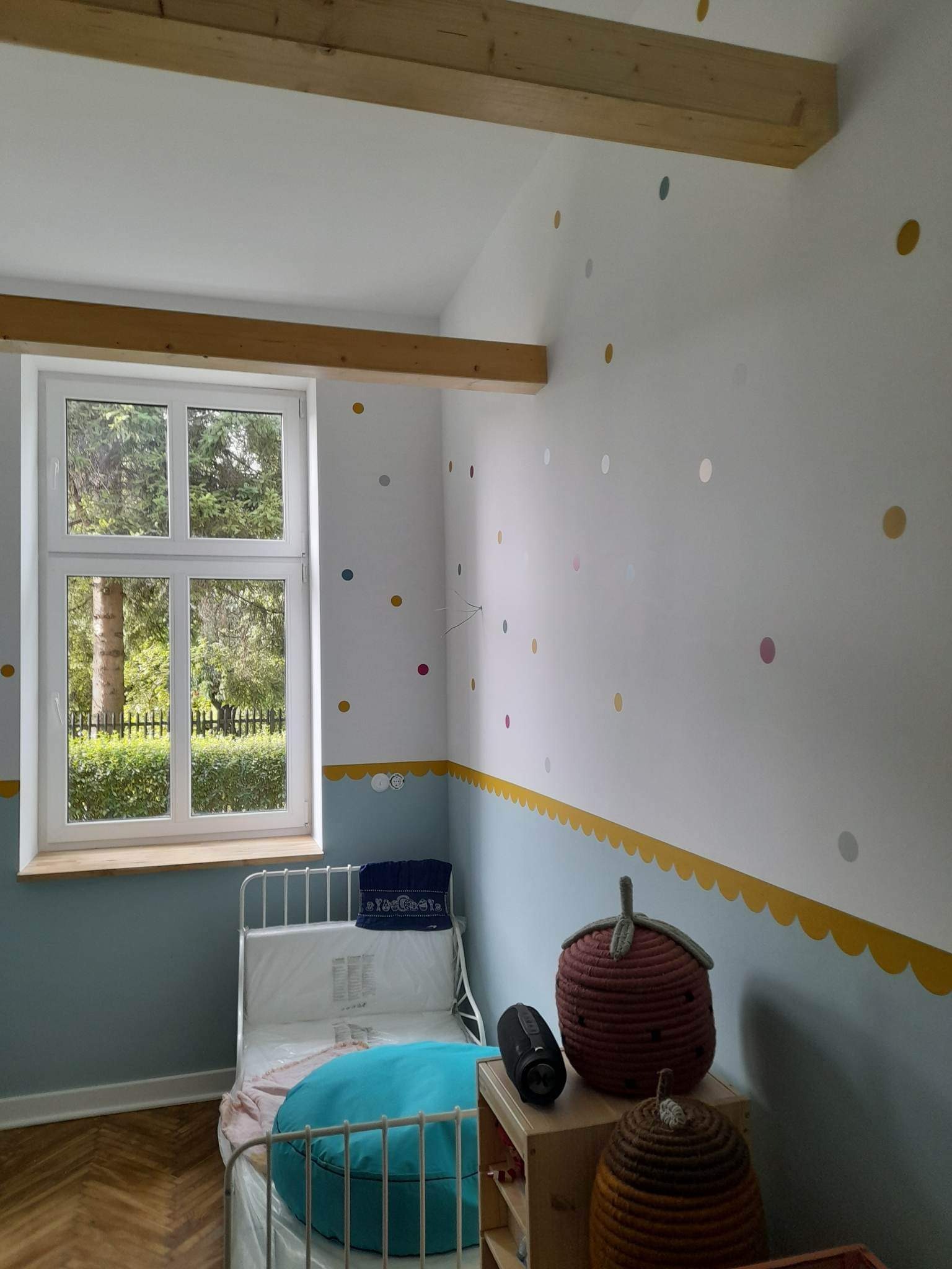 Pokój dziecięcy po remoncie: białe ściany z kolorowymi kropkami i niebieskim pasem z żółtymi falbankami, drewniane belki stropowe, białe okno z widokiem na zieleń, łóżko z turkusową poduszką...