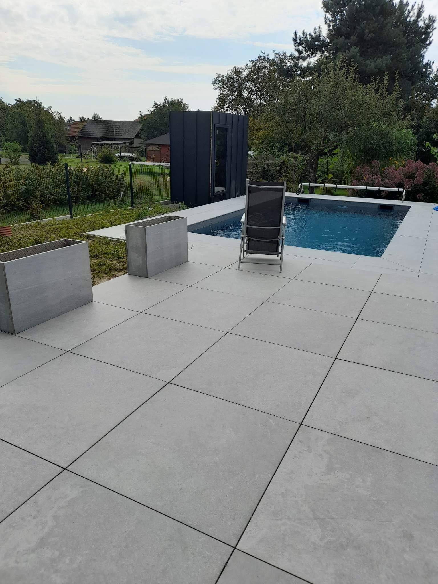 Nowoczesny taras z płyt ceramicznych w jasnoszarym odcieniu, otaczający basen z czystą, błękitną wodą. Dwa betonowe kwietniki i czarny pawilon ogrodowy uzupełniają minimalistyczny design.