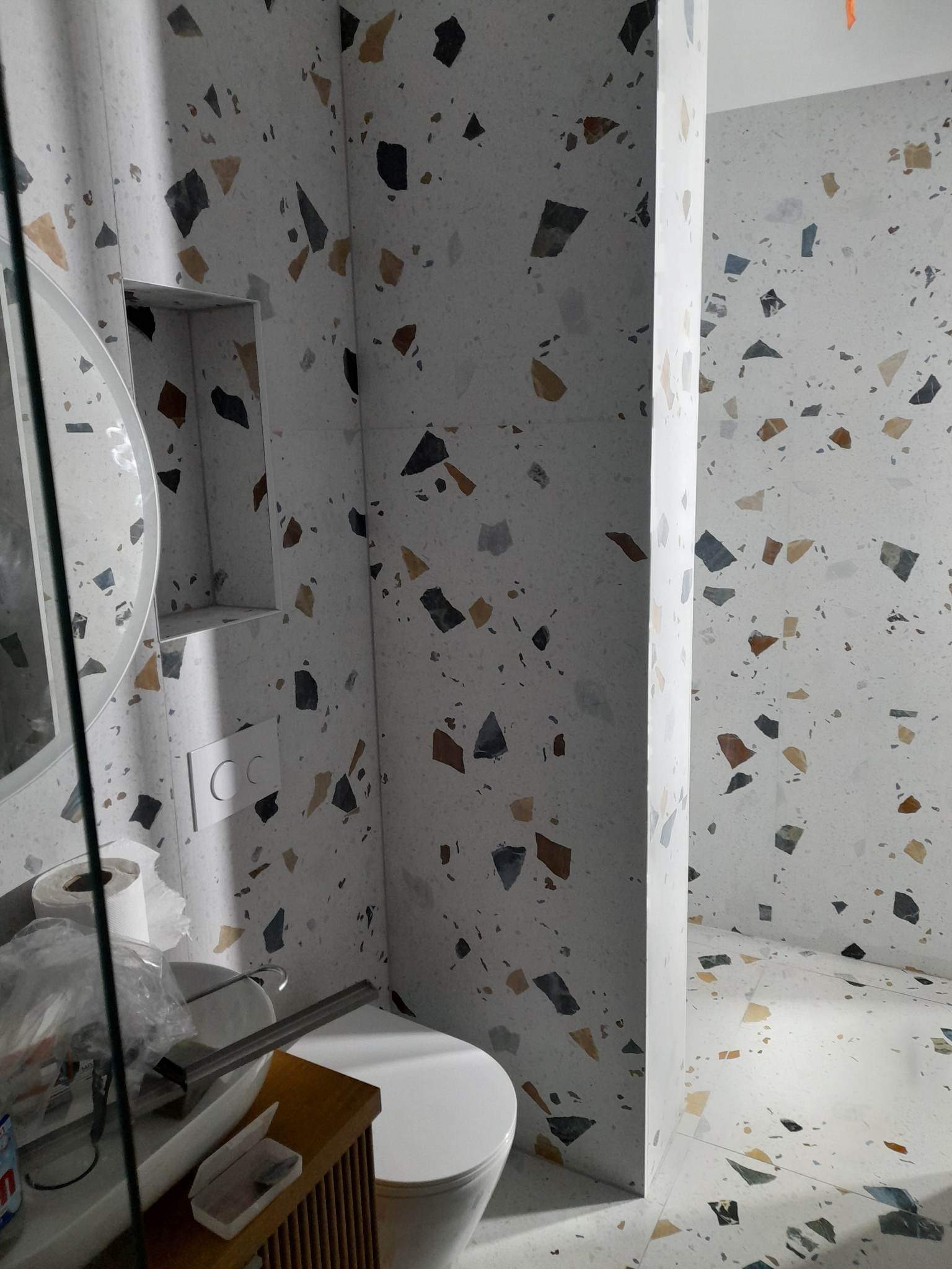 Biała łazienka wykończona płytkami imitującymi lastryko z nieregularnymi, kolorowymi fragmentami kamieni, widoczna umywalka z akcesoriami i toaleta