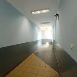 Korytarz biurowy z pomalowanymi na niebiesko ścianami w górnej części i szarymi w dolnej, z żółtą podłogą z płytek, oświetlony lampami sufitowymi i wyposażony w kamerę przemysłową.