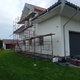 Dom jednorodzinny z białymi ścianami zewnętrznymi w trakcie prac elewacyjnych, widoczne rusztowania, zabezpieczenia folią, narzędzia i pojemnik na preparat przy ścianie.