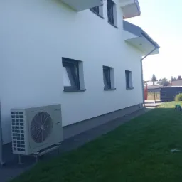 Boczny widok na dom z białą, gładką elewacją, szarymi oknami i klimatyzatorem na trawniku przed budynkiem.