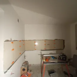 Montaż płytek ściennych w kuchni, widoczne płytki z pomarańczowymi krzyżykami dystansowymi, listwa poziomująca, oraz przecinarka do płytek na stole z czerwonymi nogami, w tle okno.