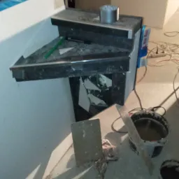 Wykonanie półki z ciemnego kamienia z widocznym uszkodzeniem okleiny bocznej, obok wiadra z zaprawą i narzędziami na zakurzonym podłożu w trakcie prac wykończeniowych.