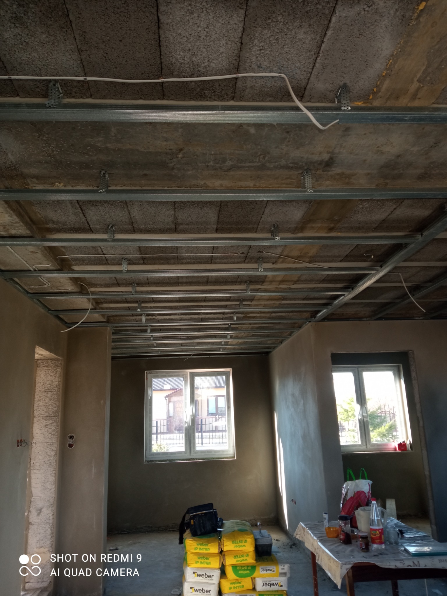 Wnętrze w trakcie prac wykończeniowych: sufit z metalową konstrukcją podwieszaną i widocznymi przewodami, ściany częściowo otynkowane, okna z widokiem na zewnątrz, na podłodze worki z zaprawą Weber...