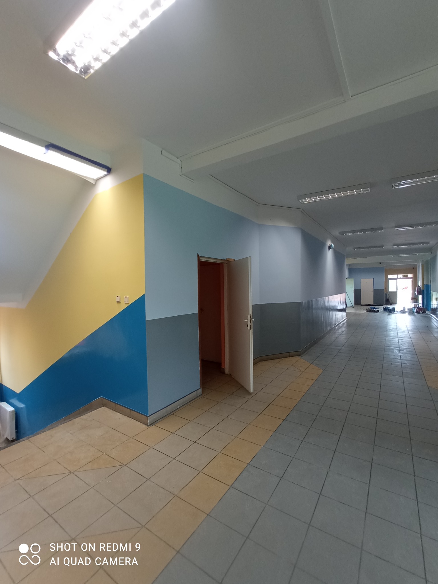 Pomalowany korytarz szkolny z kolorowymi ścianami: żółto-niebieskie schody, szaro-błękitne ściany wzdłuż korytarza, szara podłoga z beżowymi akcentami, otwarte drzwi i widoczne narzędzia malarskie...