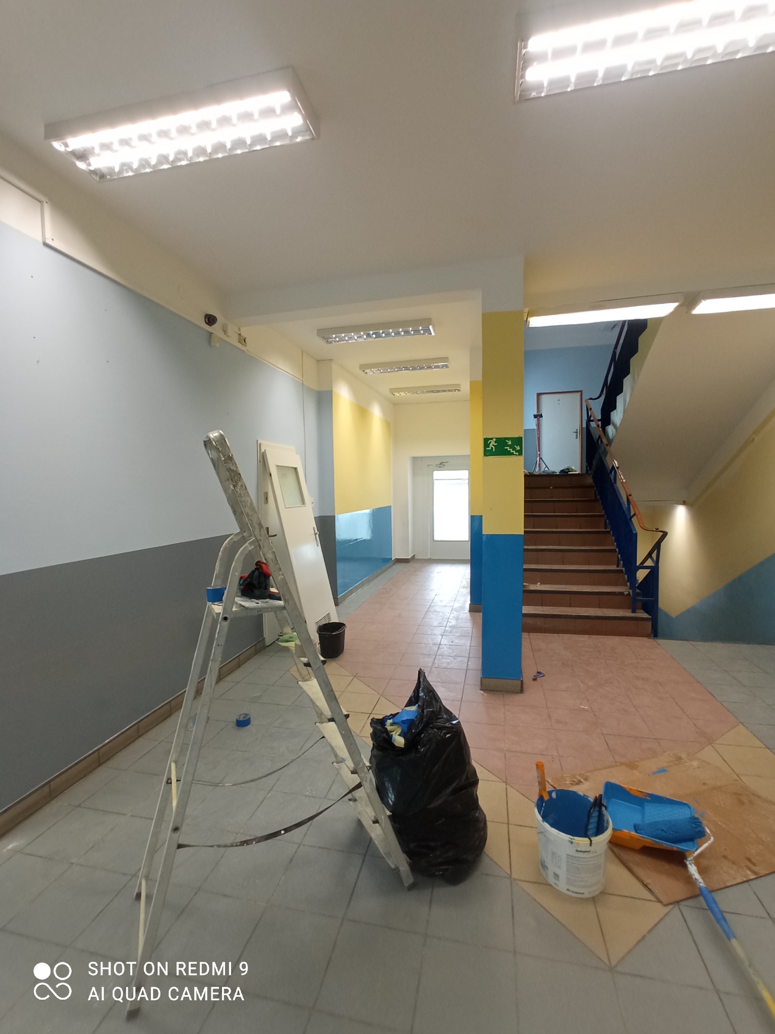 Malowanie klatki schodowej w budynku: ściany pomalowane na szaro i niebiesko, drabina przystawiona do ściany, wiadro z niebieską farbą i wałek malarski na podłodze, widoczny znak ewakuacyjny.