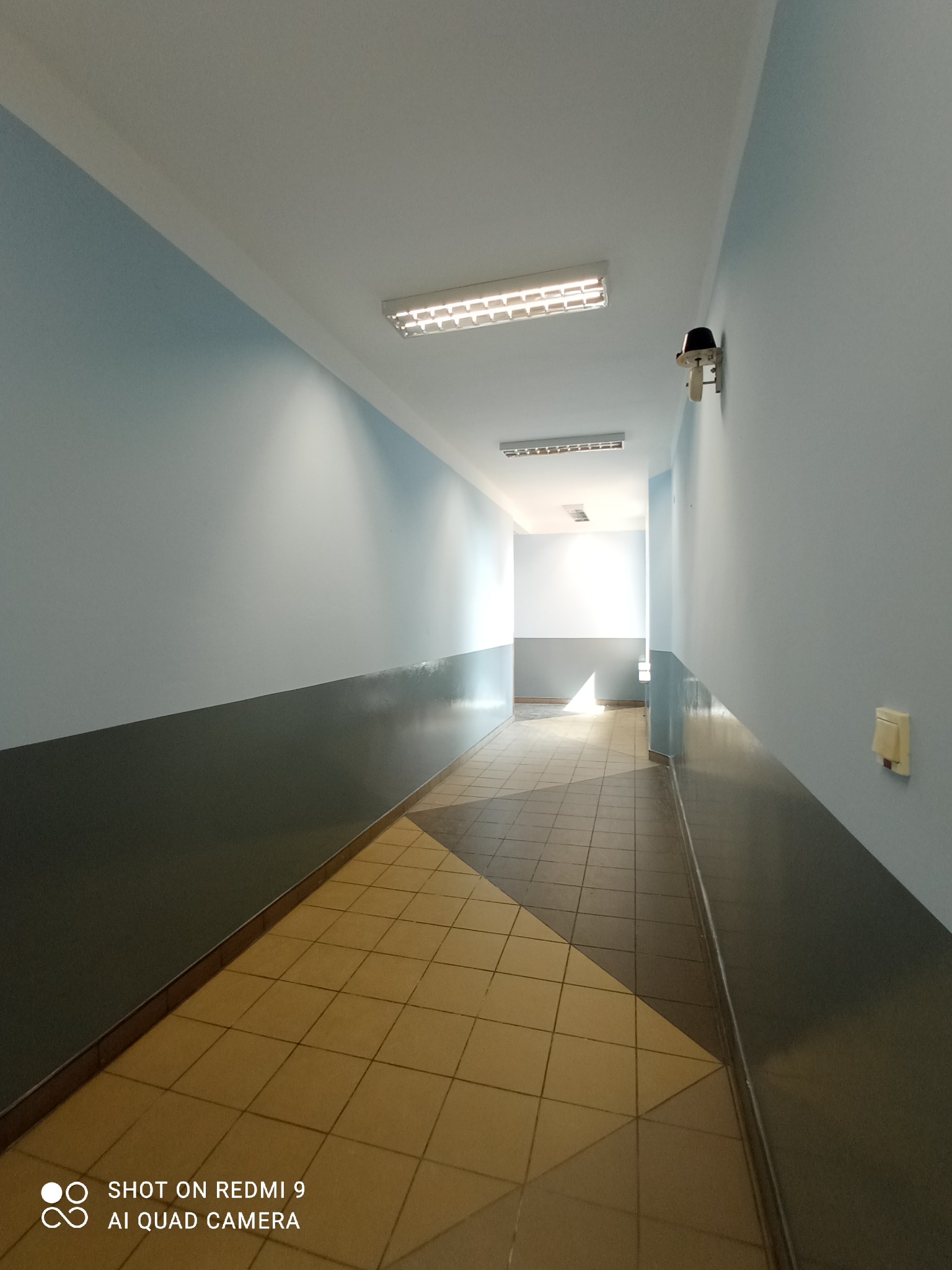Korytarz biurowy z pomalowanymi na niebiesko ścianami w górnej części i szarymi w dolnej, z żółtą podłogą z płytek, oświetlony lampami sufitowymi i wyposażony w kamerę przemysłową.