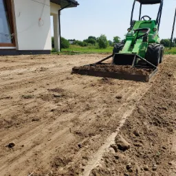 Zielona minikoparka Avant z równiarką wyrównuje teren pod przyszły ogród, widoczny fragment budynku w tle.