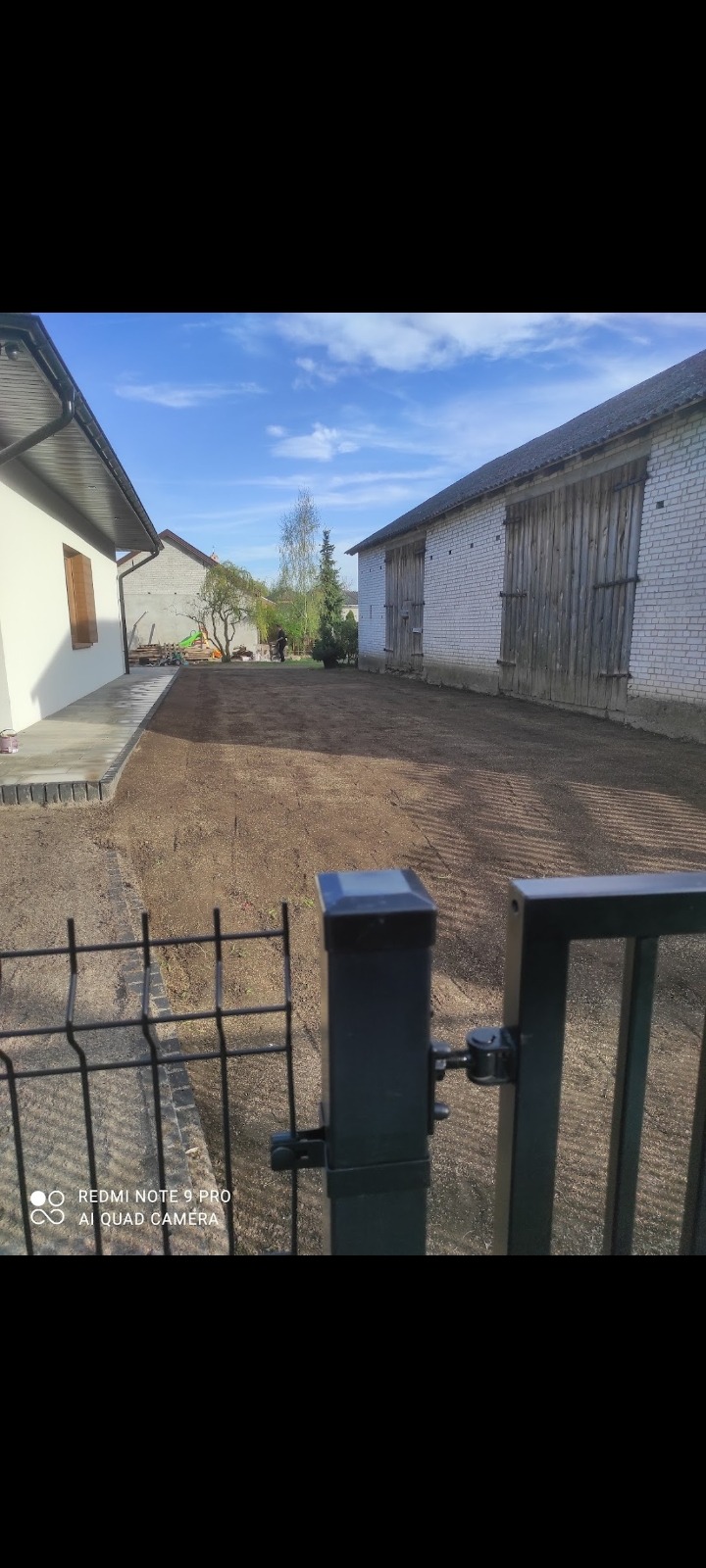 Przygotowany teren pod trawnik, widok zza metalowego ogrodzenia, budynki w tle, osoba w oddali.