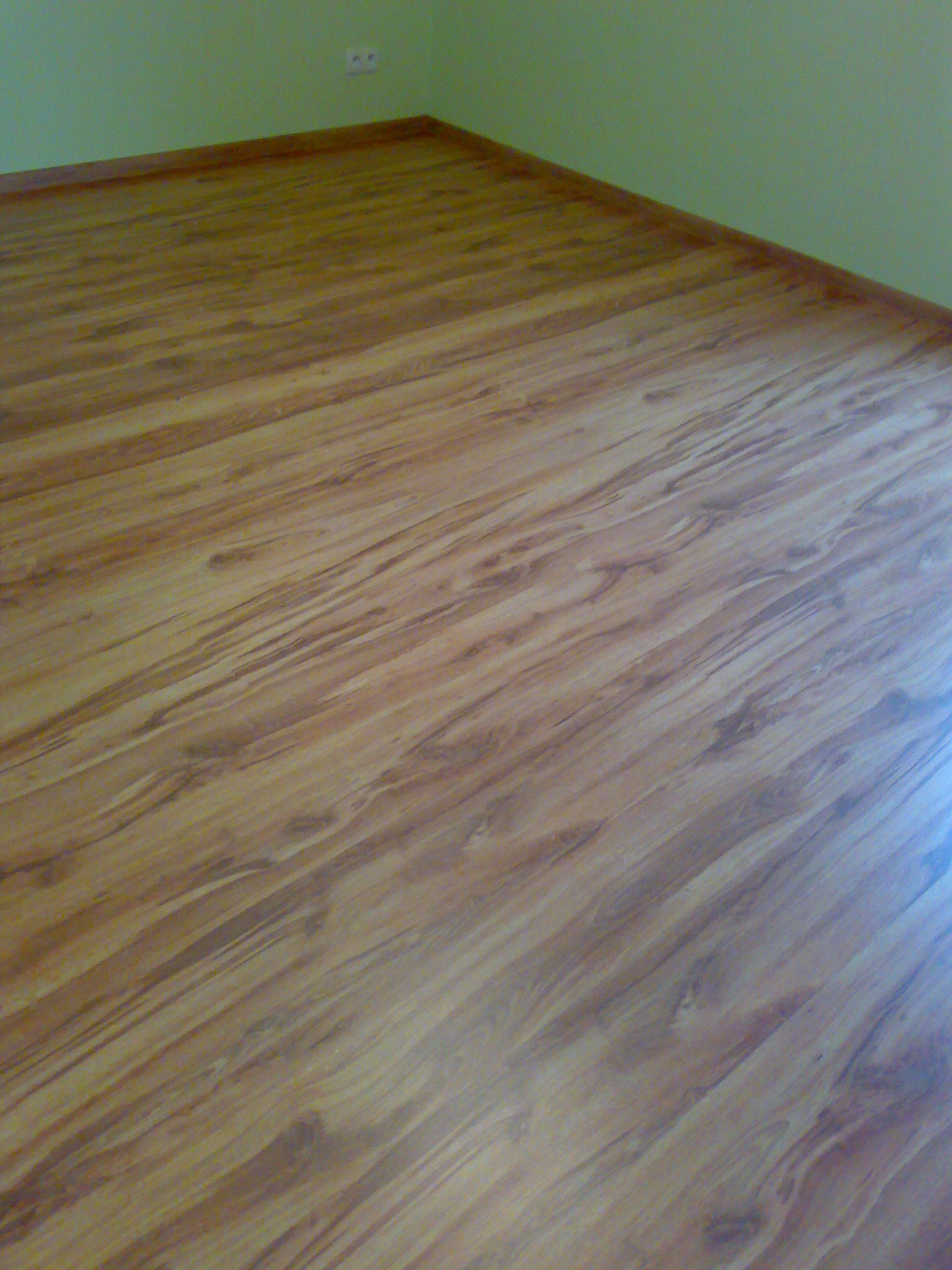 Ułożone panele podłogowe o wzorze drewna, widoczne fragmenty zielonej ściany i listwy przypodłogowe.