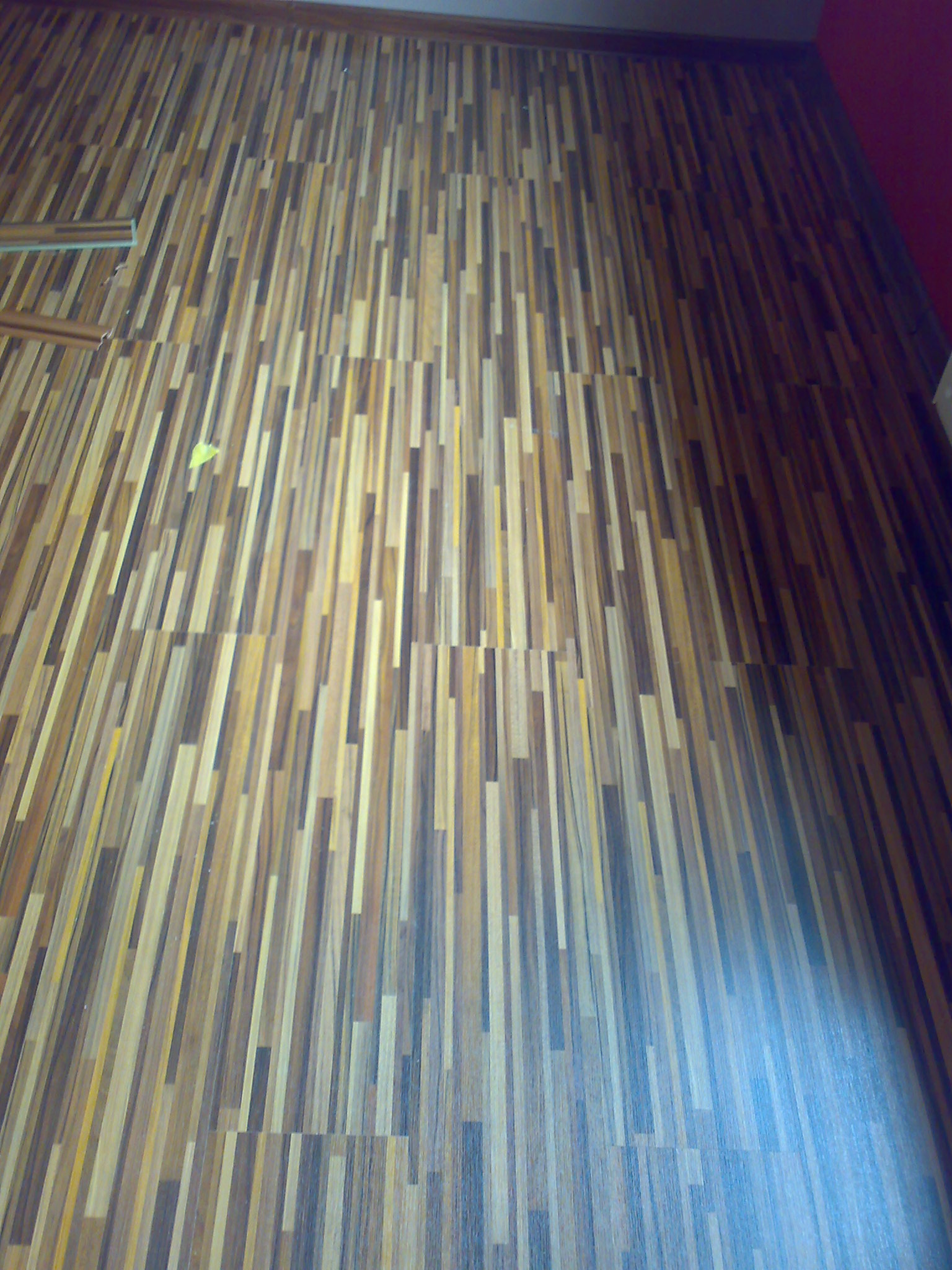 Ułożona podłoga z wąskich, różnokolorowych desek, z widocznymi fragmentami listew przypodłogowych i żółtą kartką na powierzchni.