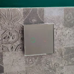 Szklany panel dotykowy włącznika światła z podświetleniem LED (zielony i czerwony) zamontowany na dekoracyjnej ścianie z motywem paisley, widoczny fragment zielonej ściany powyżej.