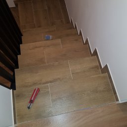 DZIKU Uslugi Remontowo-Wykonczeniowe Tomasz Gorzel - Klatka schodowa wyłożona brązowymi płytkami imitującymi drewno, z widocznym nożem do cięcia płytek, niebieskim opakowaniem po fugach i fragmentem buta roboczego w dolnej części kadru.