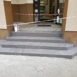 Świeżo wykafelkowane, szare schody wejściowe do budynku, zabezpieczone taśmą ostrzegawczą, z widocznymi tubami silikonu i zaprawą na pierwszym stopniu.