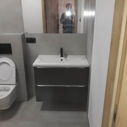 DZIKU Uslugi Remontowo-Wykonczeniowe Tomasz Gorzel - Nowoczesna łazienka w szarościach z podwieszaną szafką umywalkową i lustrem, w którym odbija się osoba robiąca zdjęcie.