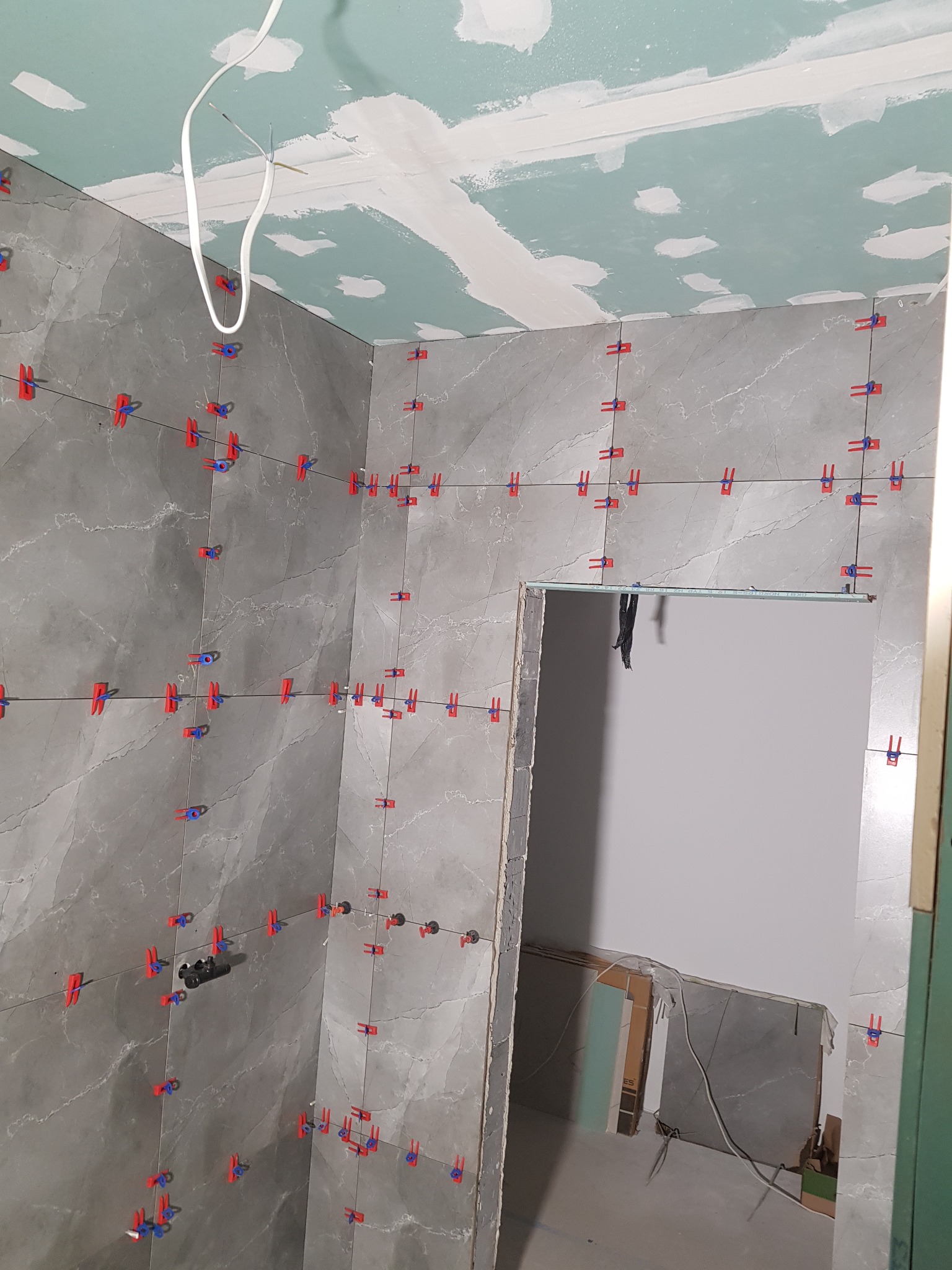 Ściany łazienki w trakcie układania płytek imitujących marmur, z użyciem systemu poziomowania płytek (klipsy i kliny), widoczne przewody elektryczne i surowy sufit z płyt gipsowo-kartonowych.
