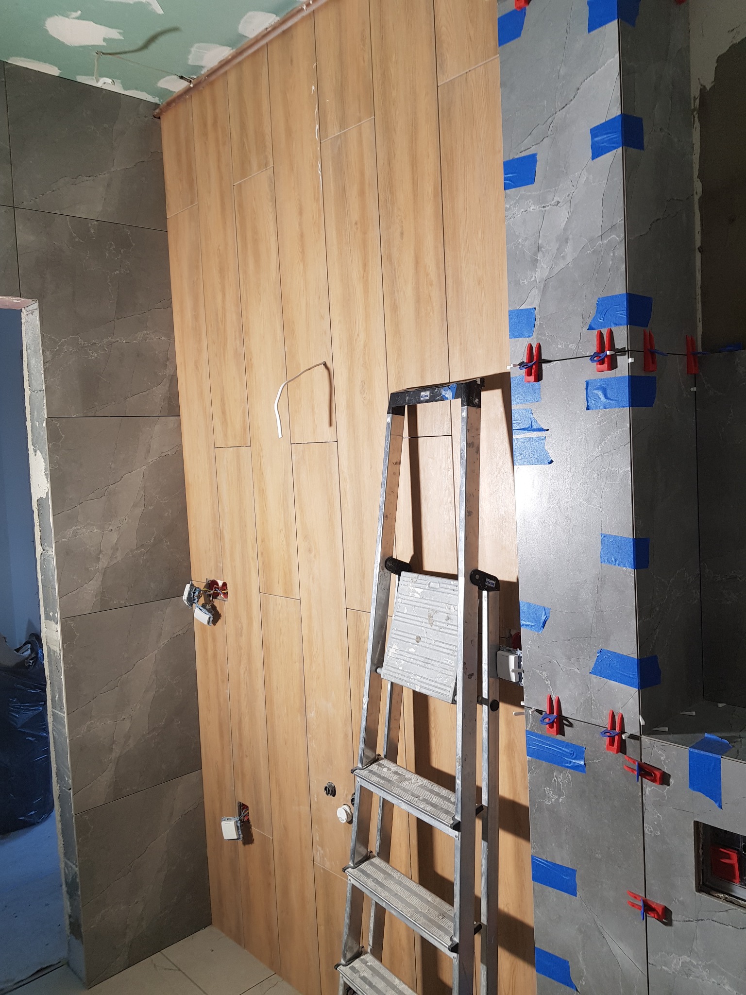 Montaż płytek imitujących drewno na ścianie z instalacją elektryczną oraz szarych płytek z niebieską taśmą i czerwonymi krzyżykami poziomującymi, widoczna drabina.