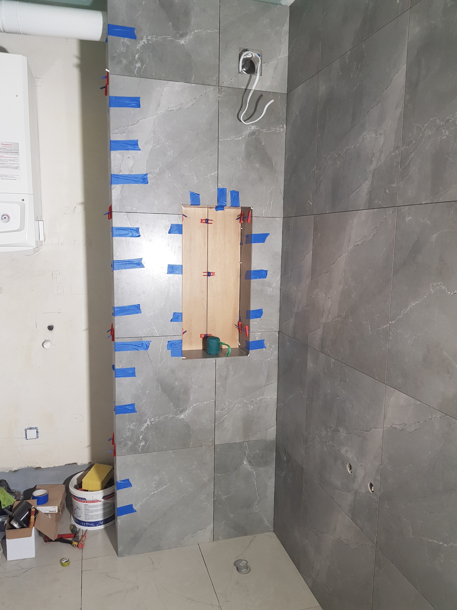 Ściana łazienki w trakcie układania szarych płytek imitujących marmur, z widocznym wnęką zabezpieczoną drewnianą ramą i niebieską taśmą, oraz fragmentem kotła gazowego i narzędziami na podłodze.