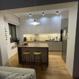 Nowoczesna kuchnia z drewnianym blatem, hokerami i białymi szafkami, oświetlona wiszącą lampą i reflektorami punktowymi, widok z salonu.