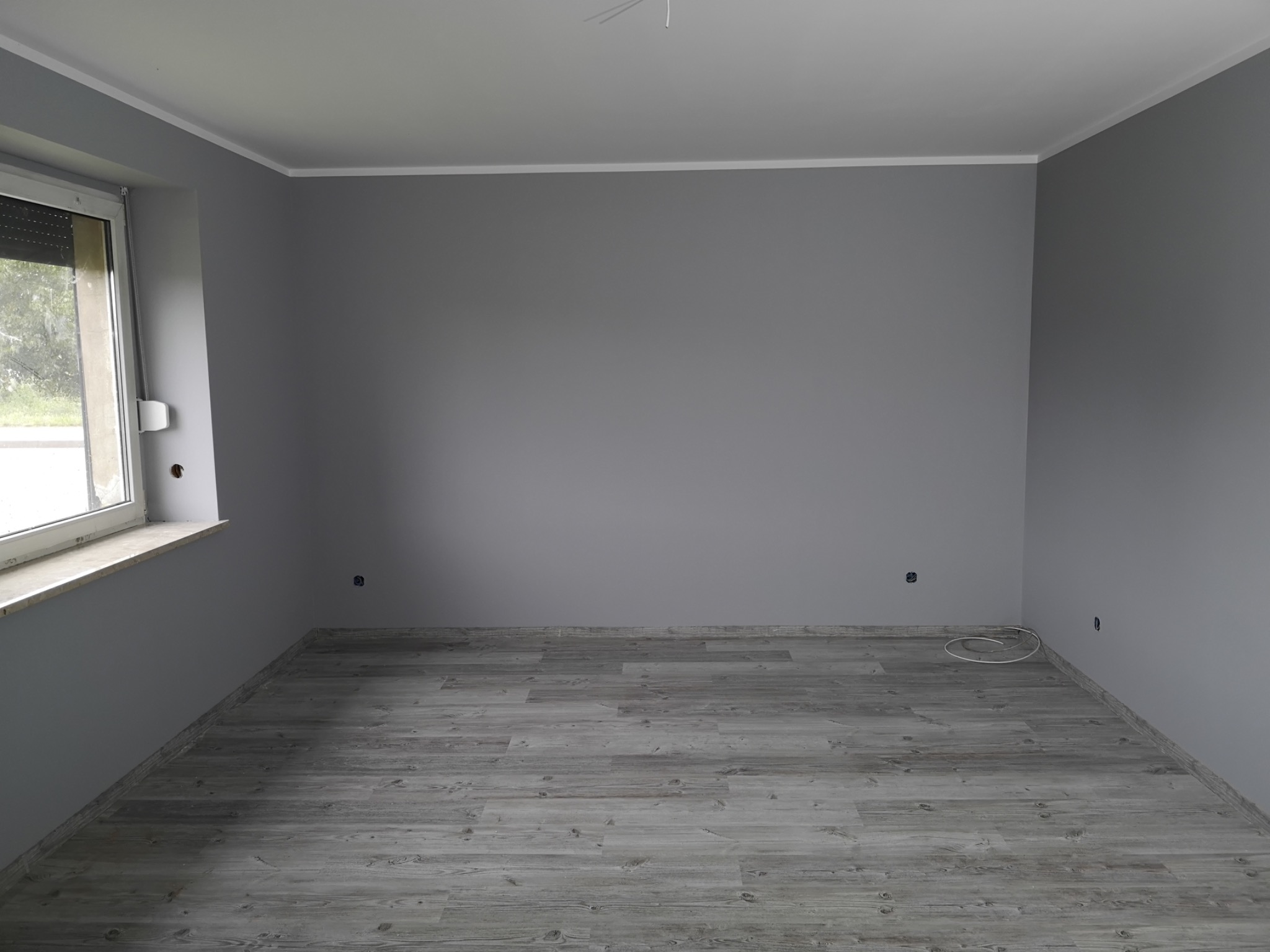 Pokój po malowaniu na szaro, z drewnopodobną podłogą i wystającym kablem przy listwie przypodłogowej.