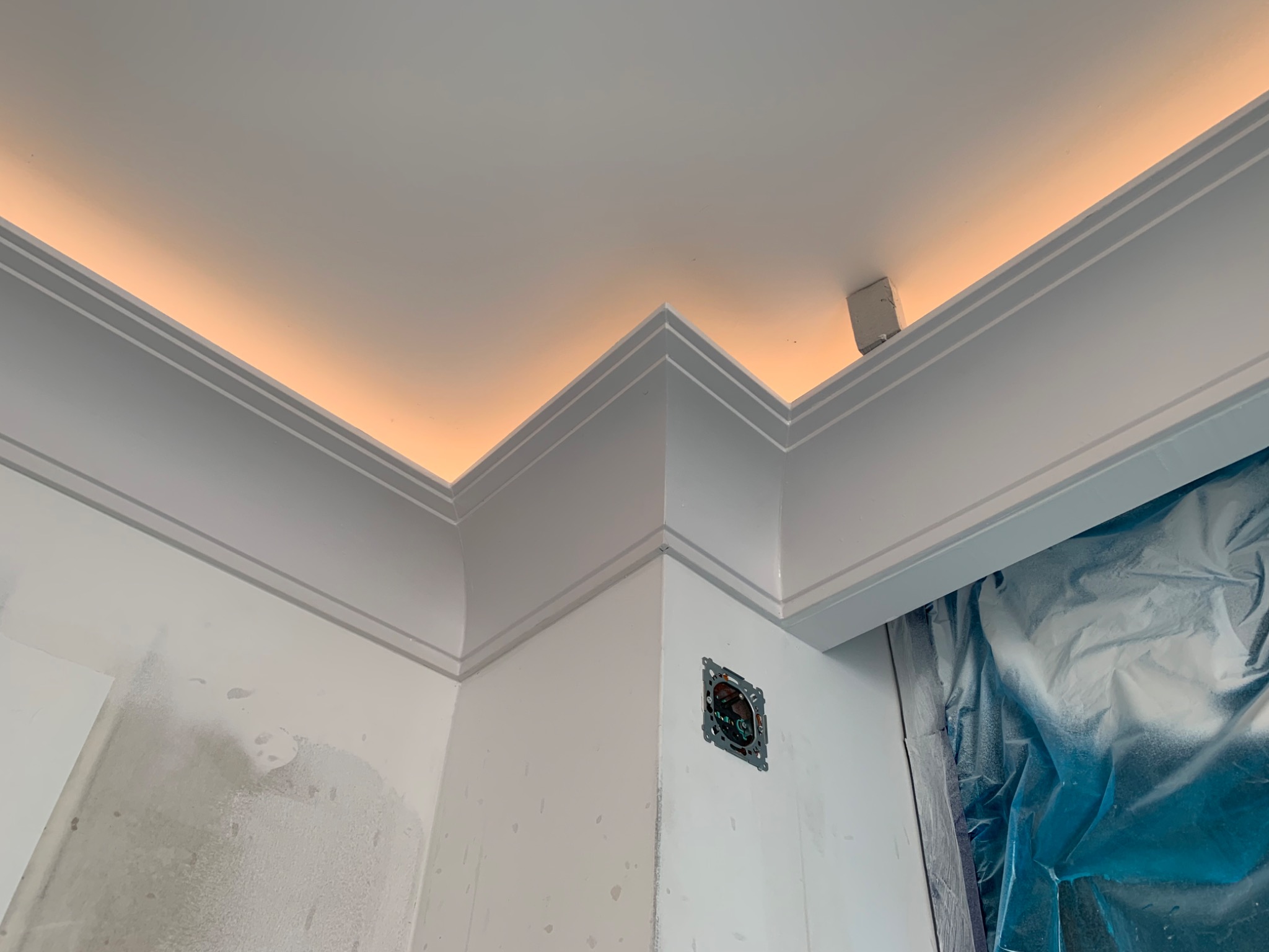 Wnętrze w trakcie prac wykończeniowych: narożnik sufitu z zamontowanym oświetleniem LED i listwami dekoracyjnymi, ściany po gładziowaniu, widoczne gniazdko elektryczne i folia ochronna.