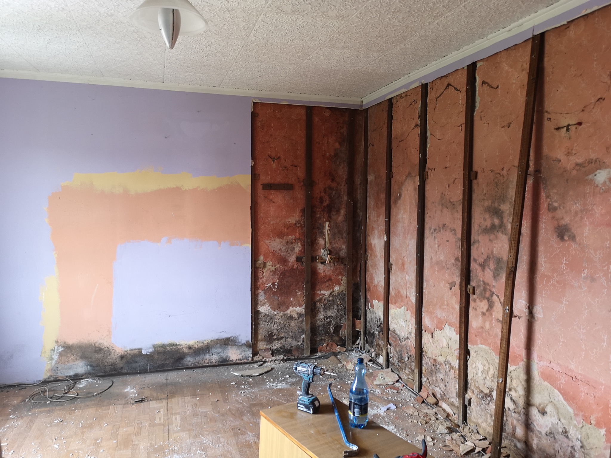 Pokój w trakcie remontu: ściany częściowo ogołocone z tapet i farby, odsłonięta konstrukcja, widoczne ślady wilgoci i pleśni, na podłodze resztki materiałów, wiertarka i butelka wody na szafce.