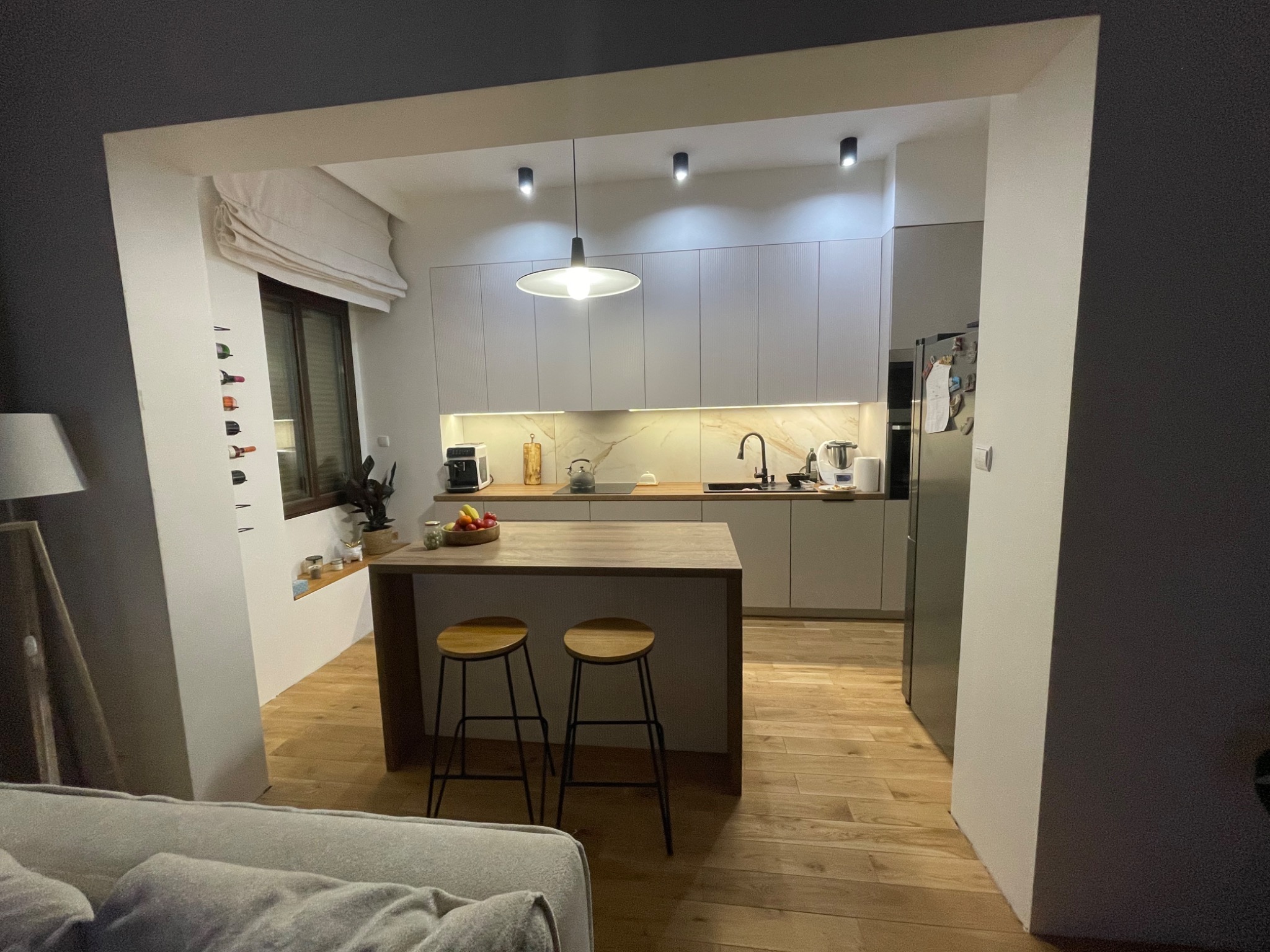 Nowoczesna kuchnia z drewnianym blatem, hokerami i białymi szafkami, oświetlona wiszącą lampą i reflektorami punktowymi, widok z salonu.