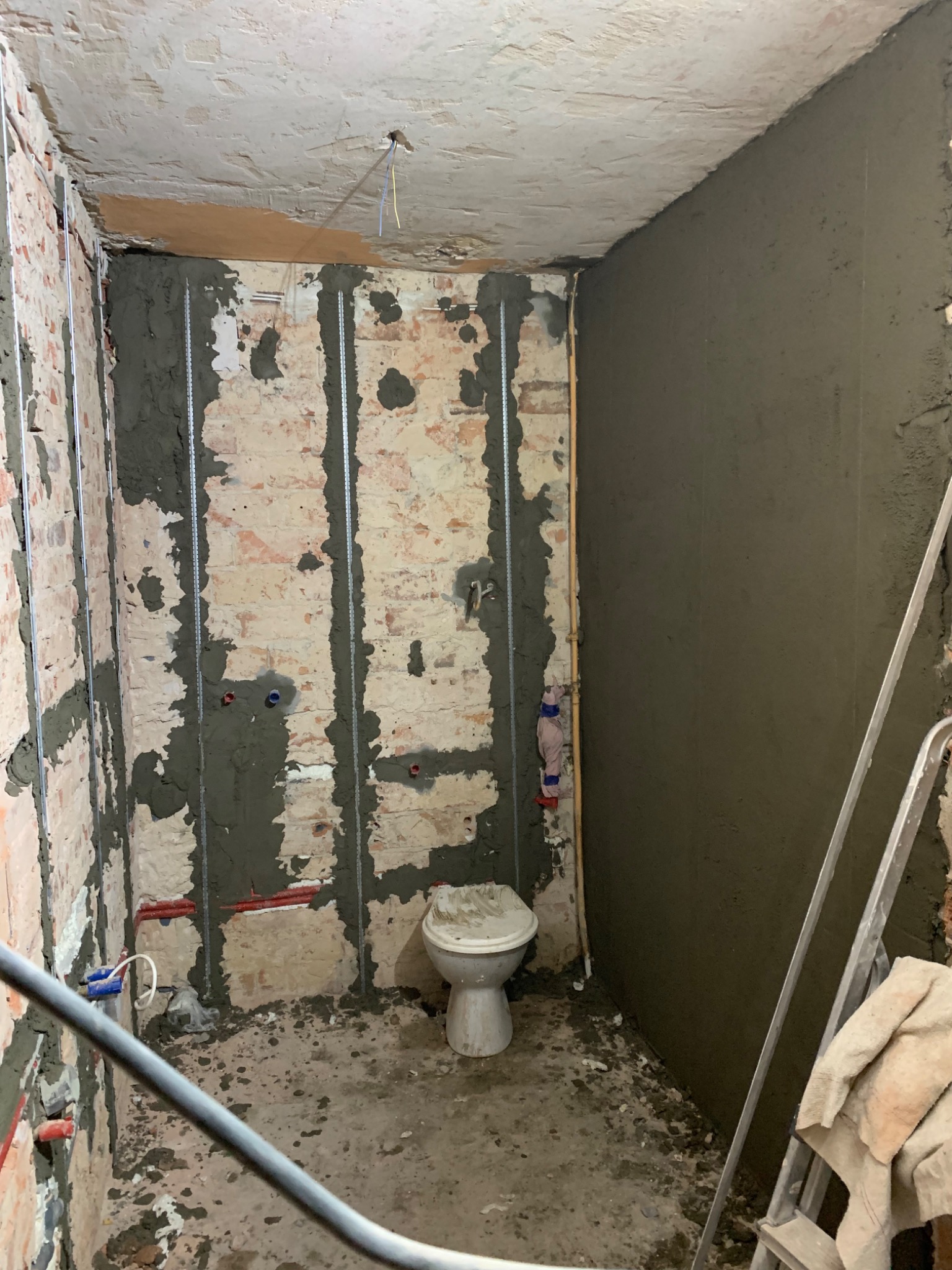 Remontowana łazienka z częściowo otynkowaną ścianą, widoczne profile pionowe na ścianach z cegły, stara toaleta i drabina oparta o ścianę.