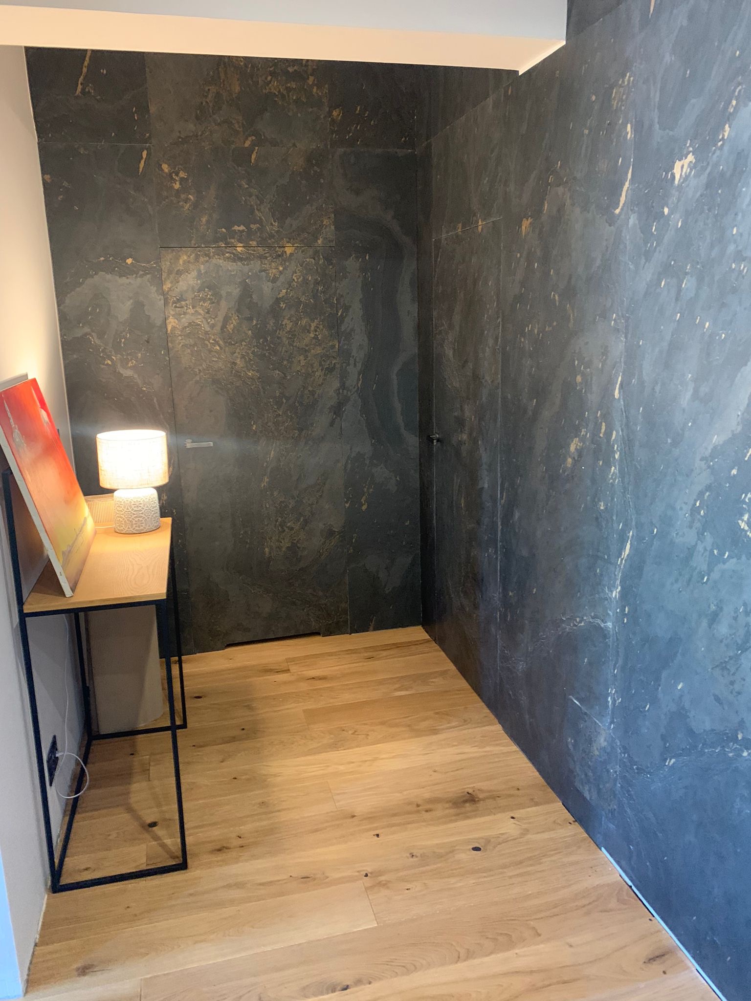 Wykończona ściana w łazience z ciemnych, wzorzystych płytek imitujących kamień, ukryte drzwi, drewniana podłoga i stolik z lampką.