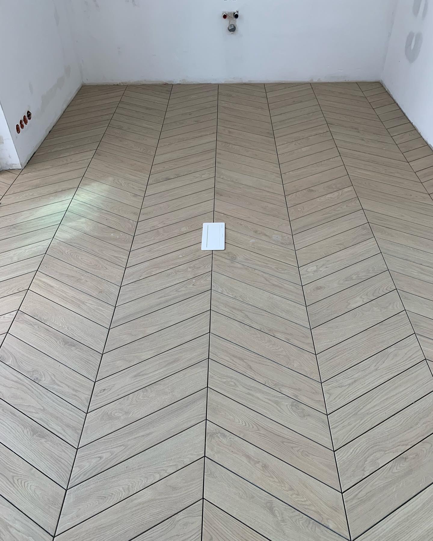 Ułożona w jodełkę podłoga z płytek imitujących drewno, biała ściana w tle z widocznymi puszkami elektrycznymi i rurami.