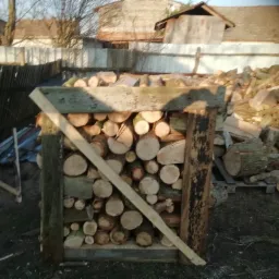 Klatka z surowego drewna wypełniona pociętymi okrąglakami, w tle sterta drewna i zabudowania gospodarcze, widoczne prowizoryczne wzmocnienia konstrukcji.