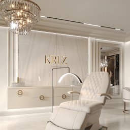 Załuczkowski Studio Interior Architecture - Elegancki gabinet kosmetyczny z beżowym fotelem do zabiegów, marmurową ścianą z podświetlanym logo 'KRUZ Aesthetics', kryształowym żyrandolem i minimalistycznymi szafkami.