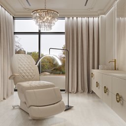 Załuczkowski Studio Interior Architecture - Jasne wnętrze z fotelem kosmetycznym z regulowanym podnóżkiem, nowoczesną lampą łukową, kryształowym żyrandolem i szafką z dekoracyjnymi uchwytami na tle okna z widokiem na jesienne drzewa.