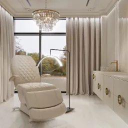 Jasne wnętrze z fotelem kosmetycznym z regulowanym podnóżkiem, nowoczesną lampą łukową, kryształowym żyrandolem i szafką z dekoracyjnymi uchwytami na tle okna z widokiem na jesienne drzewa.